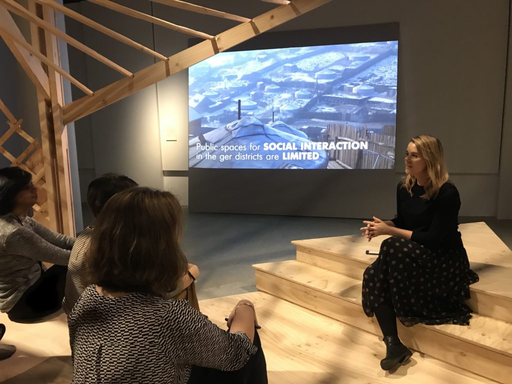 Woman sitting on wooden steps giving a talk to a group of people with presentation slides on the wall behind.